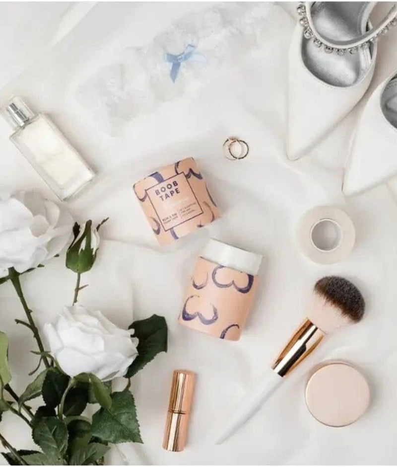 Makeup products including boob tape, a bottle, tubes, and brushes on a light surface with flowers and jewellery.