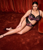Model wearing Elomi Morgan Underwired Bra and High Leg Brief Set in Dark Romance lounging on red carpet  against red chair with red curtain backdrop